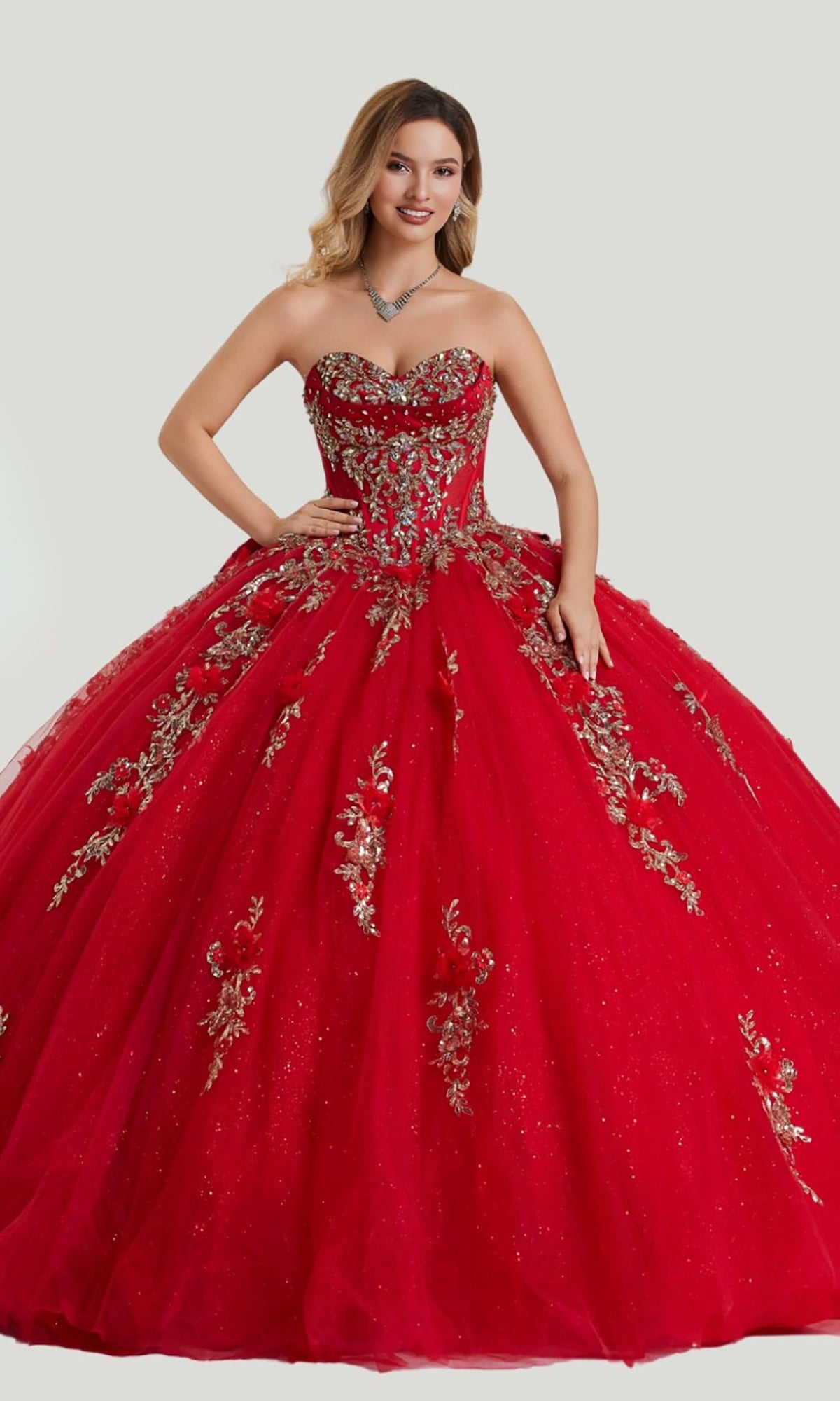 A woman stands against a light background wearing the Quinceanera Dress 26414 by Stella Couture, a strapless red ball gown embellished with gold appliques and paired with a jeweled necklace.